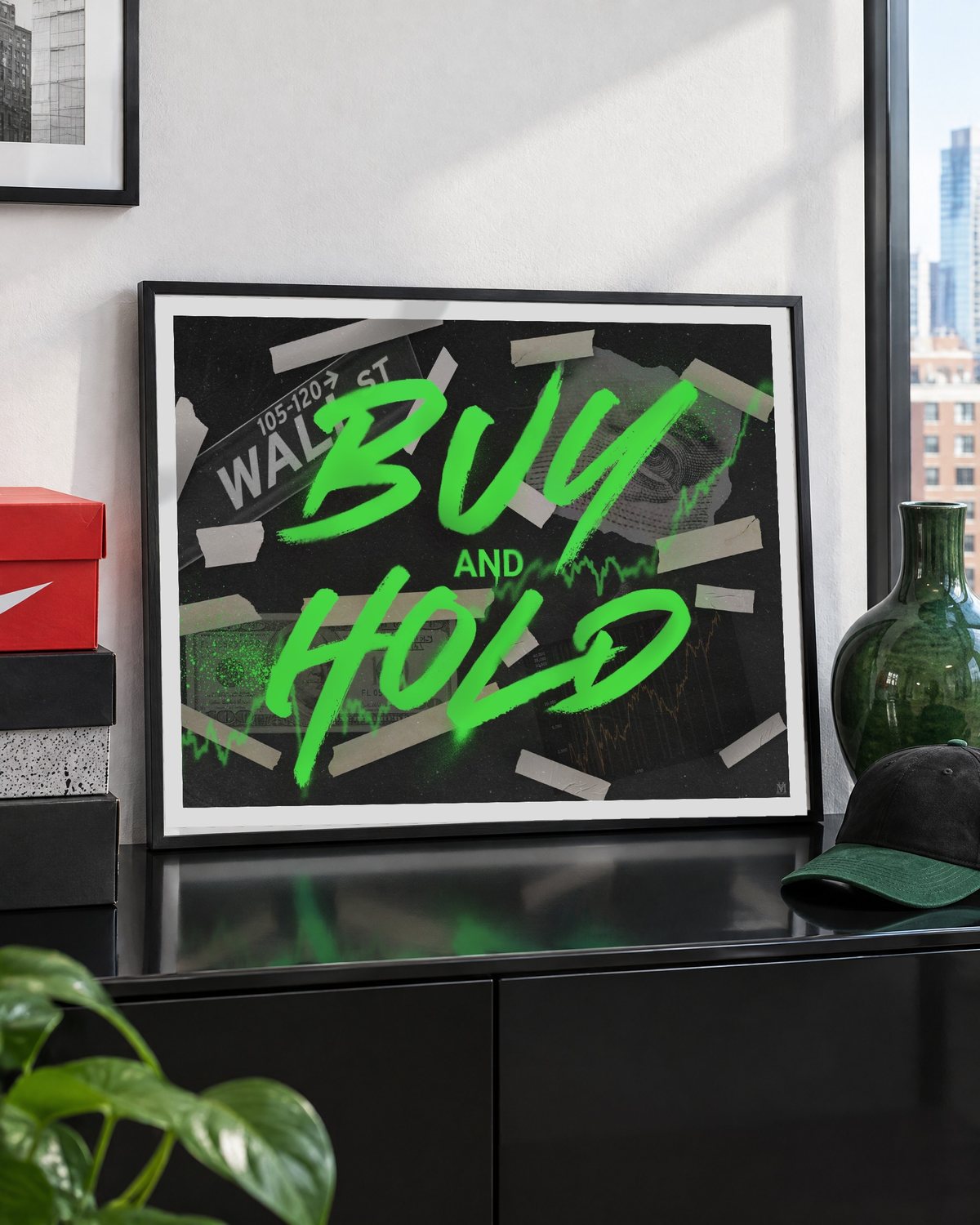 Das gerahmte Poster auf einem schwarzen Sideboard an eine weiße Wand gelehnt, daneben Schuhkartons, eine grüne Vase und eine Kappe